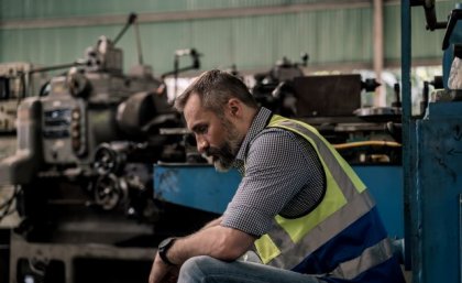 A bearded man in a short and high vis vest sits with his head bowed in a factory.